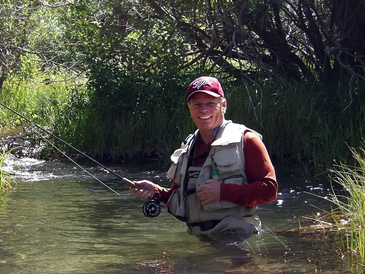 Bill Ward standing waist-deep in a clear stream, holding a fly rod