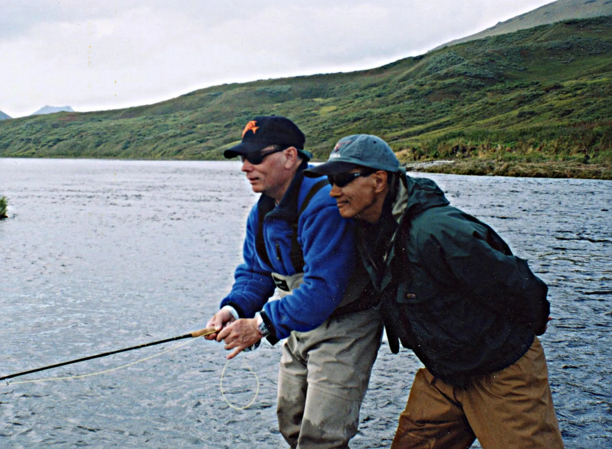 Two anglers on a riverbank below green hills, one casting a fly rod while the other watches the cast